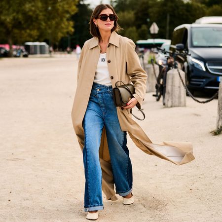 woman wears white top jeans and white shoes while wearing a trench coat over it and an olive green handbag tucked under her arm