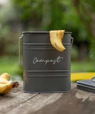 A dark gray compost bin with the letters 'compost' on it with a banana skin hanging out of it and a banana next to it, on a dark gay wooden table with a backyard scene behind it