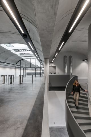 LEFT of the image The entrance of the building with grey high arched ceiling, cream walls, and a grey floor. Low glass and steel barriers that further leads into the building. RIGHT of the image shows a woman walking down a grey curved staircase.
