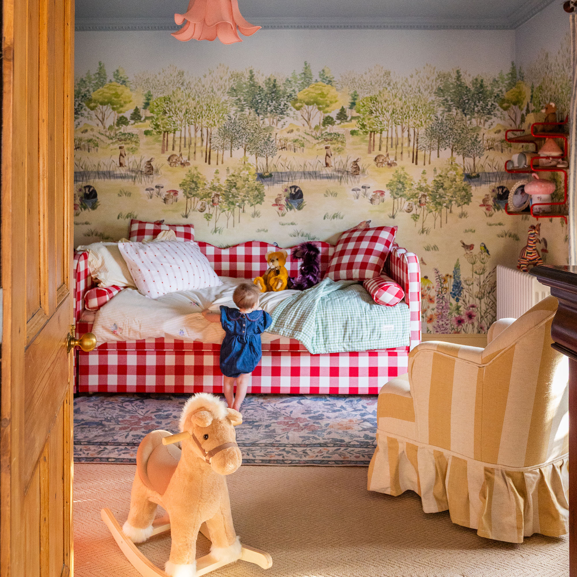 Child's bedroom with forest wall mural, yellow striped armchair, floral rug, rocking horse and red gingham daybed