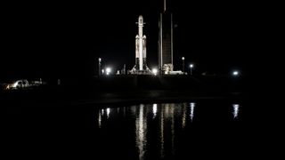 a white rocket sits on its launch pad at night, with a small lake in the foreground.