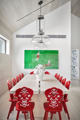 Red folksy chairs encircle a pale wood dining table backdropped by a bright green framed piece of artwork.