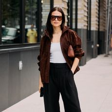 woman wears brown jacket white tank top black pants with a brown handbag tucked under her arm