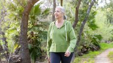 woman in a green hoodie walking on a path in a forest setting. she's looking just off camera. 