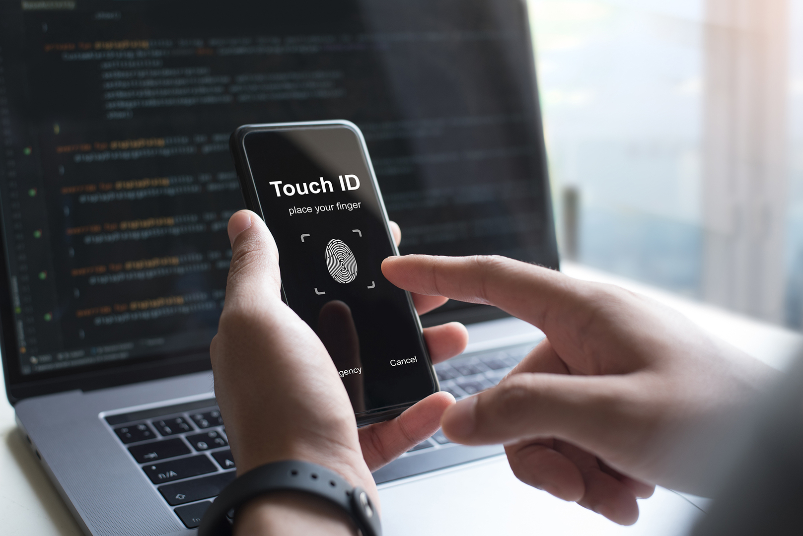jemand nutzt touch ID auf seinem Smartphone, im Hintergrund steht ein Laptop auf einem Schreibtisch