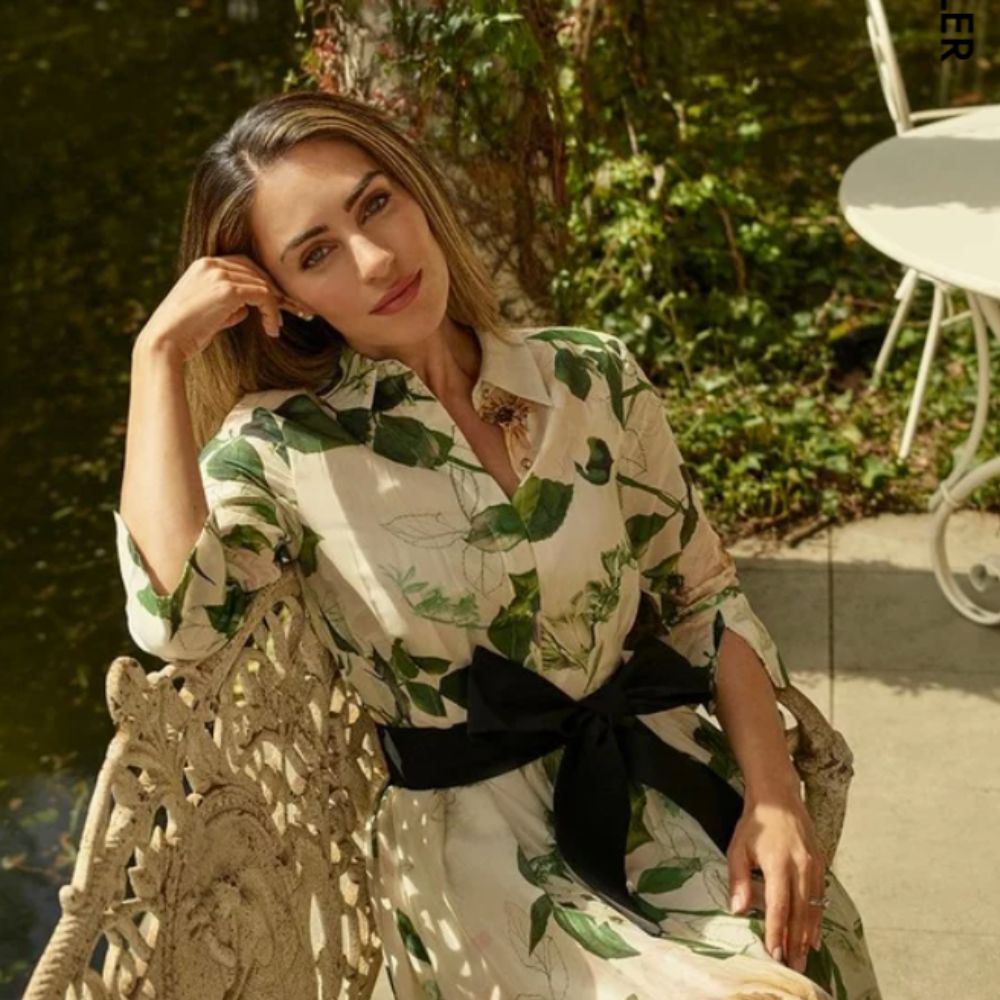 Woman staring at camera, sat on garden bench wearing a Karen Millen white dress with green bow and leaves on it.