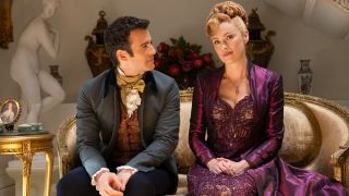 (L to R) Luke Thompson as Benedict Bridgerton, Hannah New as Lady Tilley Arnold talking while sitting on a couch.