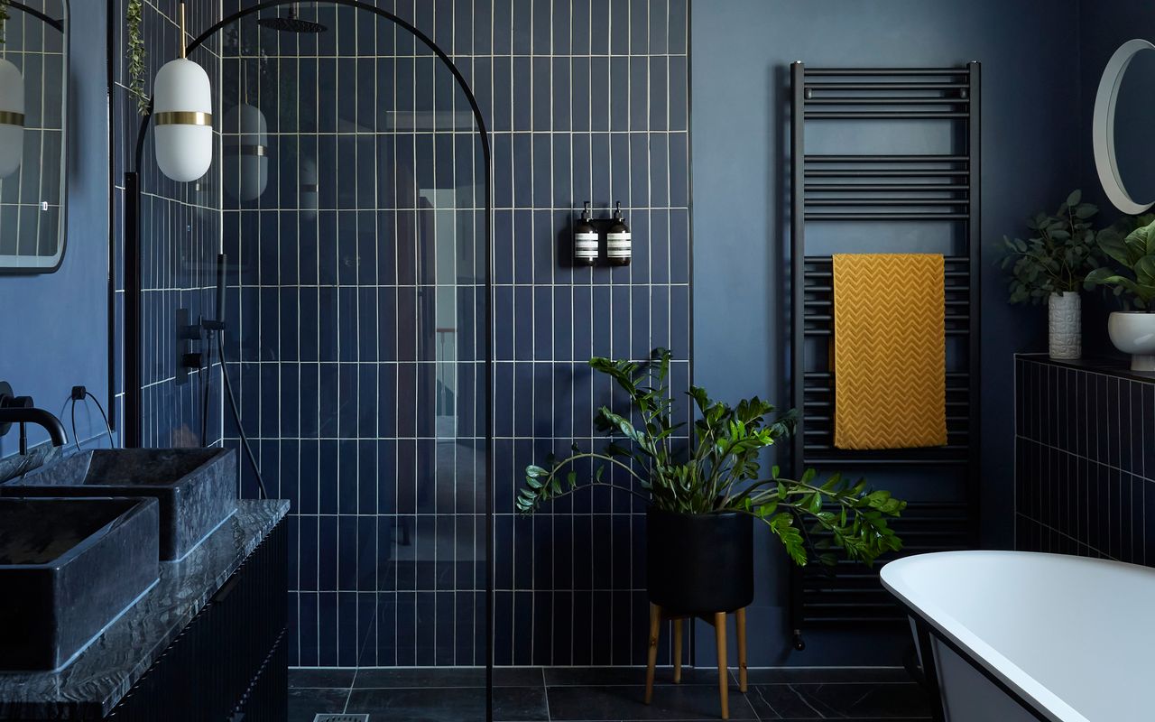 A dark navy ensuite bathroom with rectangular wall tiling, glass arch-shaped shower door, black metal heated towel rail with mustard yellow chevron patterned towel, circular mirror and white bathtub