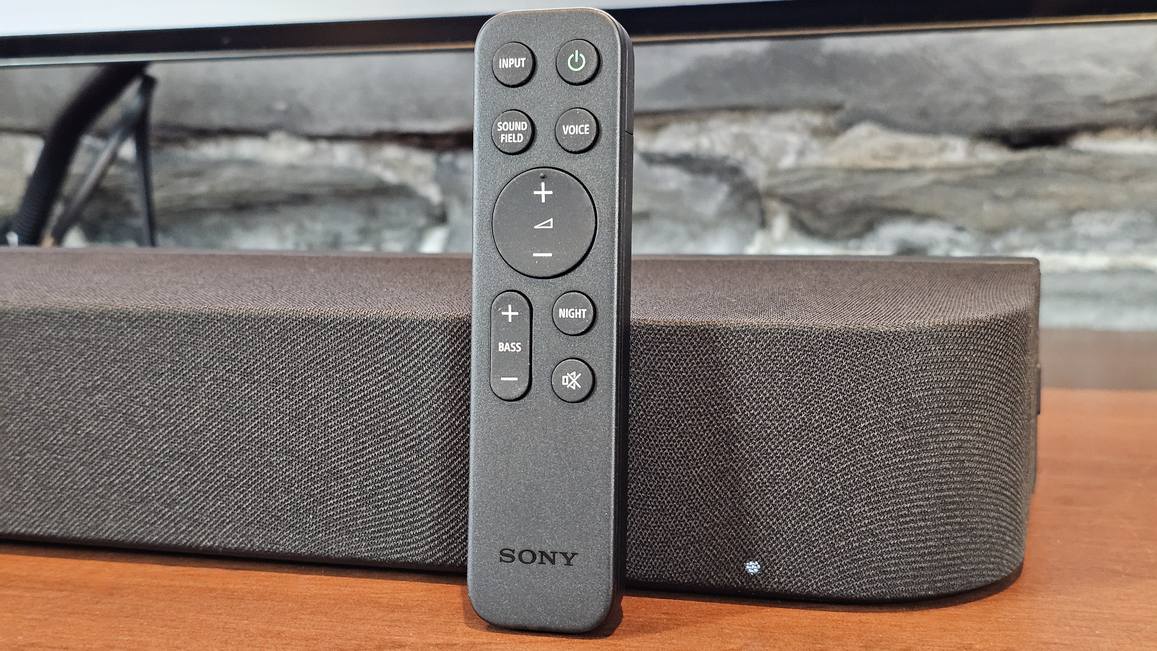 The Sony Bravia Theater Bar 7 pictured with its remote on a wooden surface underneath a TV.