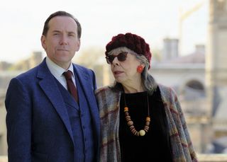 Professor T (Ben Miller) and his mother Adelaide (Frances de la Tour) stand with the Cambridge skyline in the distance behind them. She is wearing sunglasses and looking at him while he looks into the camera