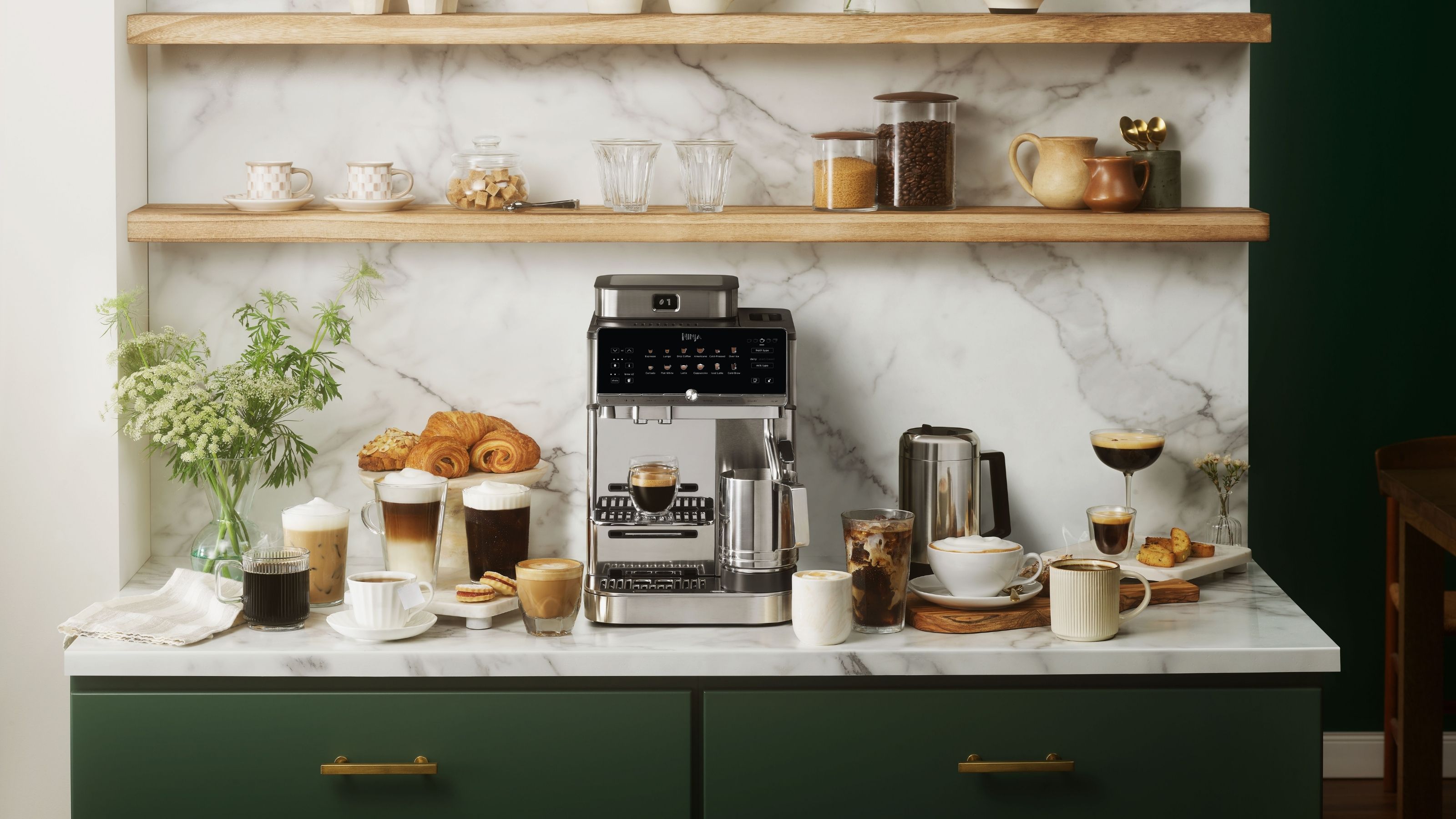 Image of a silver, Ninja coffee machine on a white marble countertop with forest green cabinetry. There are a multitude of various coffee drinks on the counter, as well as pastries. 