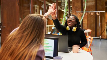 Two women share a high five and big smiles while sitting at their desks.