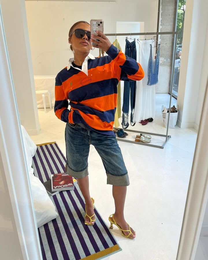 A woman standing wearing jean baggy shorts, a orange and blue polo, and yellow heels.