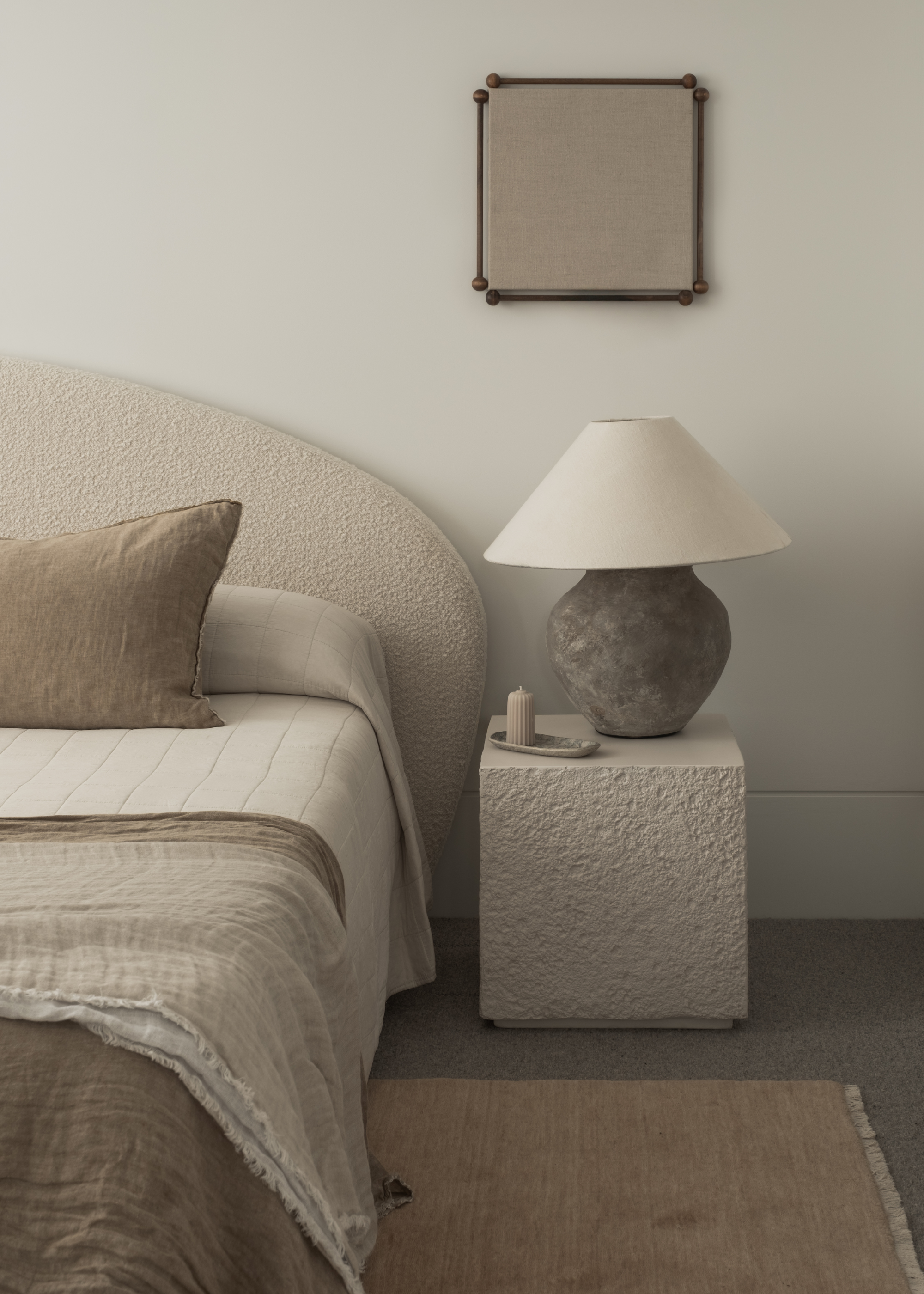 A white bedroom with a square mirror, a stone side table with a lamp, a tray with a candle, a boucle headboard, a rug, and a rustic bedscape