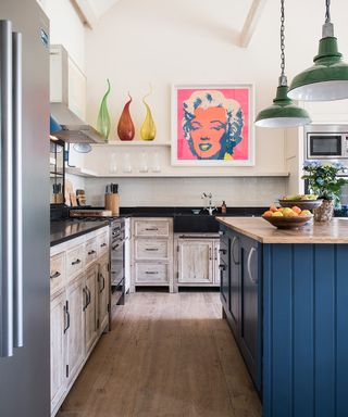 An example of kitchen extension ideas showing a kitchen with white cabinets and blue island below green pendant lamps and in front of a Marilyn Monroe artwork