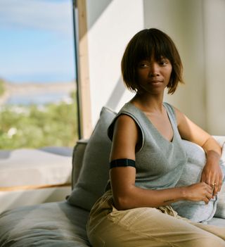 A woman with a bob haircut sits on a couch in front of a window. She wears a Visible armband on her upper arm