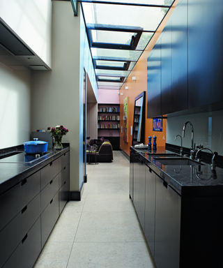 A galley kitchen with black cabinets and drawers leading to an orange wall and black bookcase