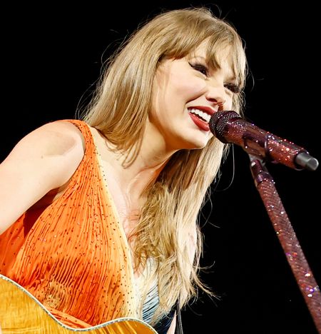 Taylor Swift wearing an orange fringed dress singing into a red microphone and holding a guitar on stage