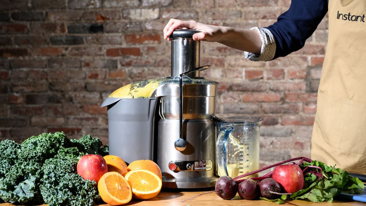 The Breville Juice Fountain Compact BJE200XL on a wooden countertop with a brick wall behind it and lots of fruits and vegetables in front of it