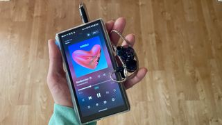 FiiO M23 player in a woman's hand, above a wooden table, playing Mogwai, Gunna and Fontaines DC