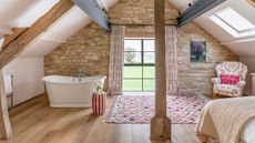An attic bedroom space with wooden flooring, exposed beams, and an exposed brick statement wall. To the right of the room is a bed with a pale peach blanket on it, and to the left is a large white bathtub. There's also a large wooden beam running up the center of the room, with a patterned pink and white rug behind it, and a patterned pink and white upholstered armchair to the left of it.