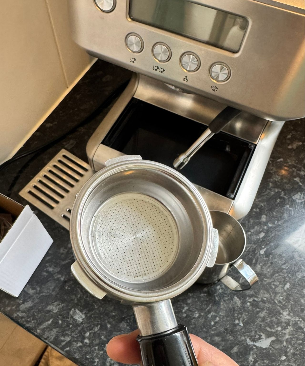Holding a portafilter above the Casabrews ULTRA Espresso Machine and its drip tray and milk jug, on black countertop.