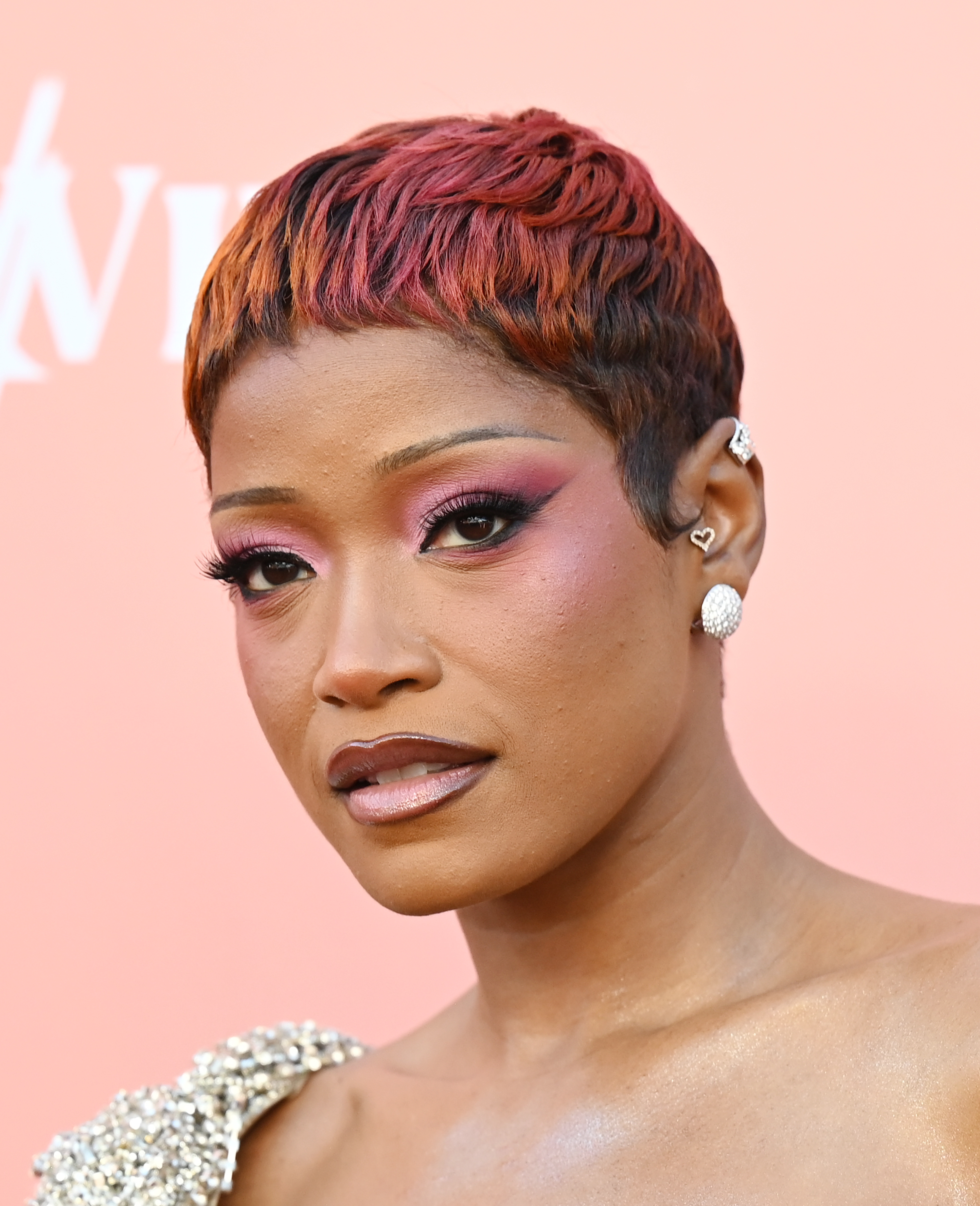 a black woman with a short pixie cut wears pink eye shadow and black eyeliner while posing in front of a peach background