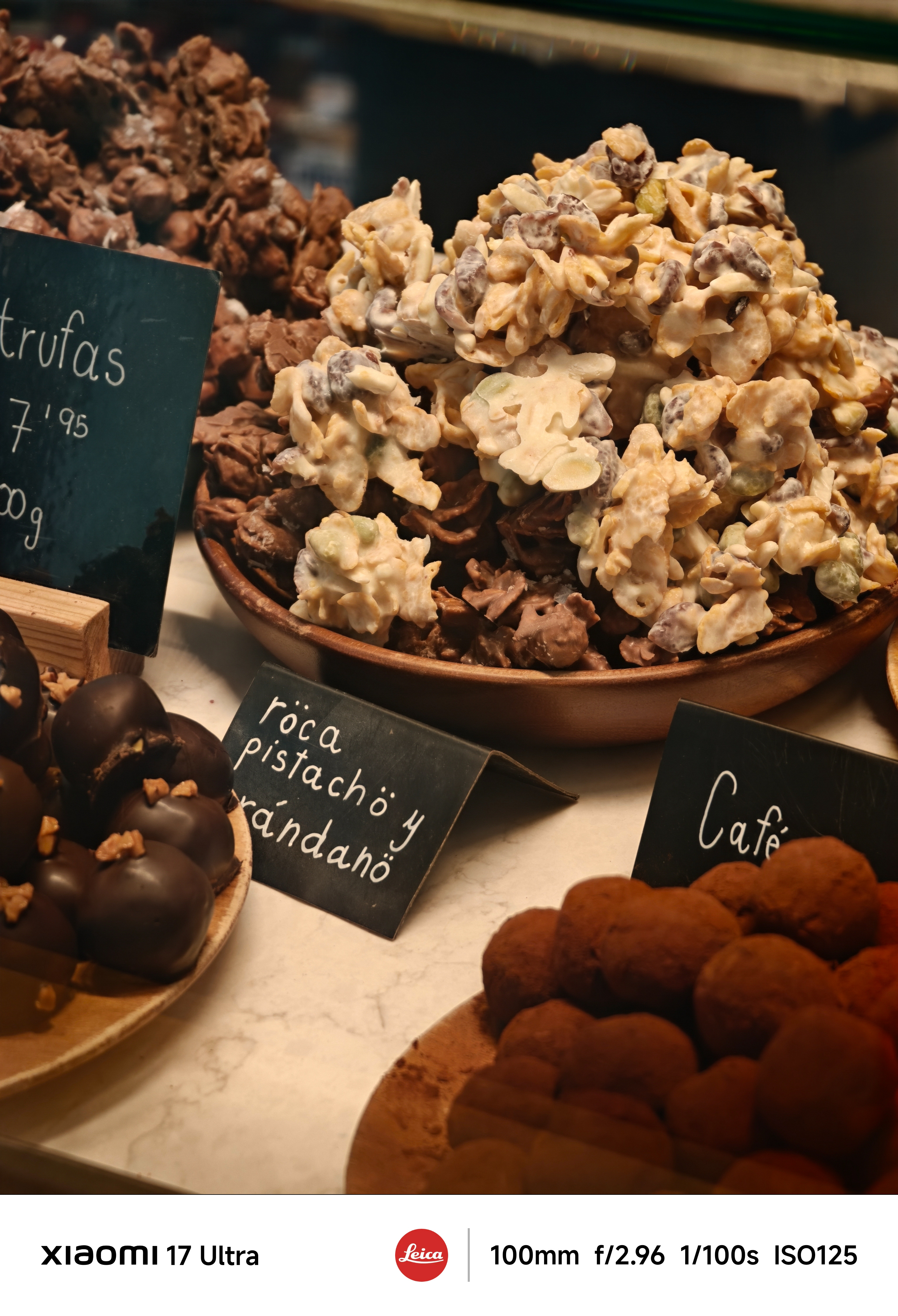 Close-up of assorted chocolates and nut clusters displayed on wooden trays behind glass, handwritten price tags visible.