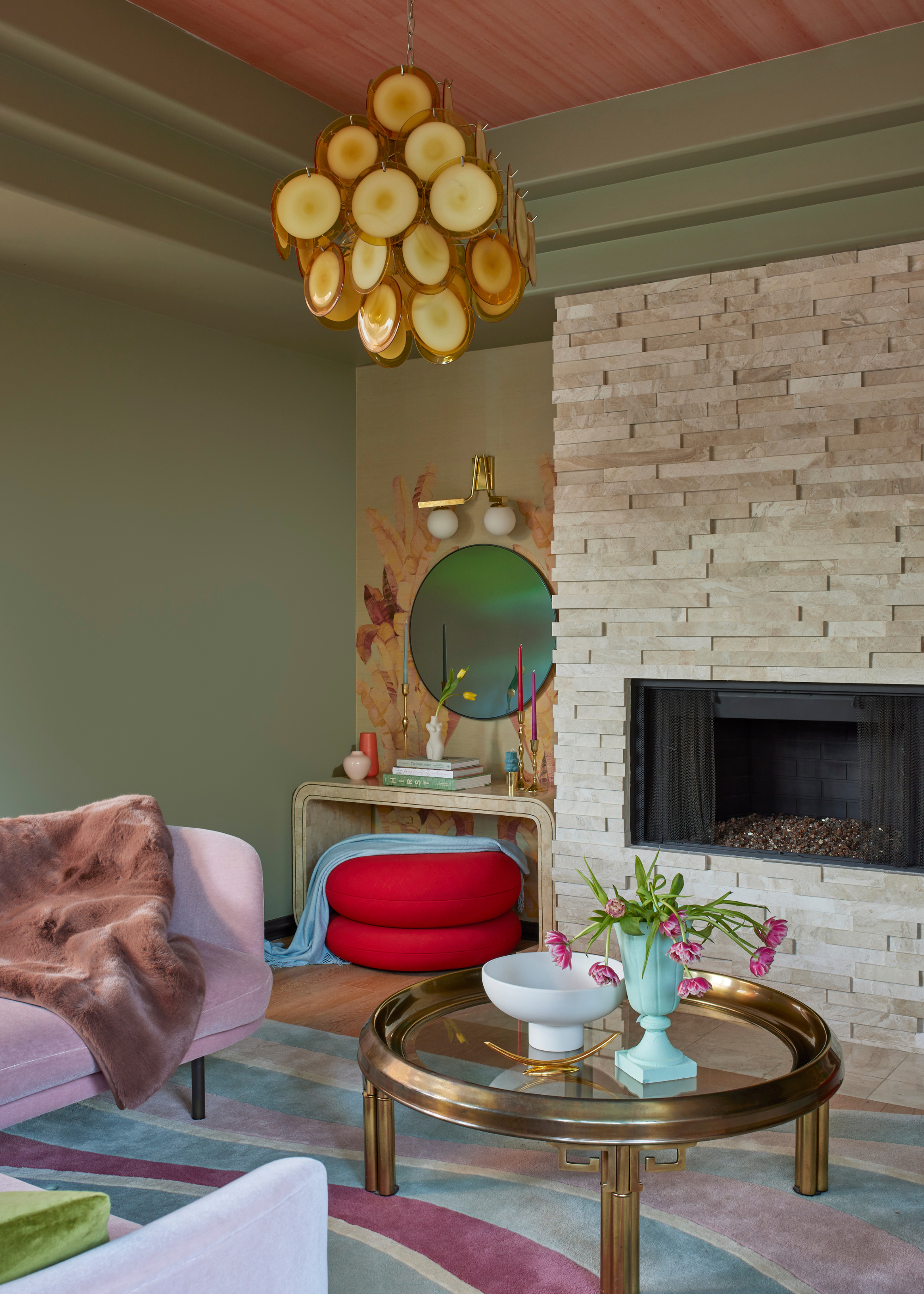A colorful living room with muted green walls, a wallpapered nook with a curved table, a wall mirror and overhead lighting near a red stool with a blue throw. A stone wall with a fireplace, a wavy striped area rug, a brass glass coffee table with a vase of tulips and a pedestal bowl and a pink couch with a plush throw.