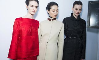 Three female models, one in a red dress, one in a beige coat with belt detail at the neck, and one in a black coat and belt