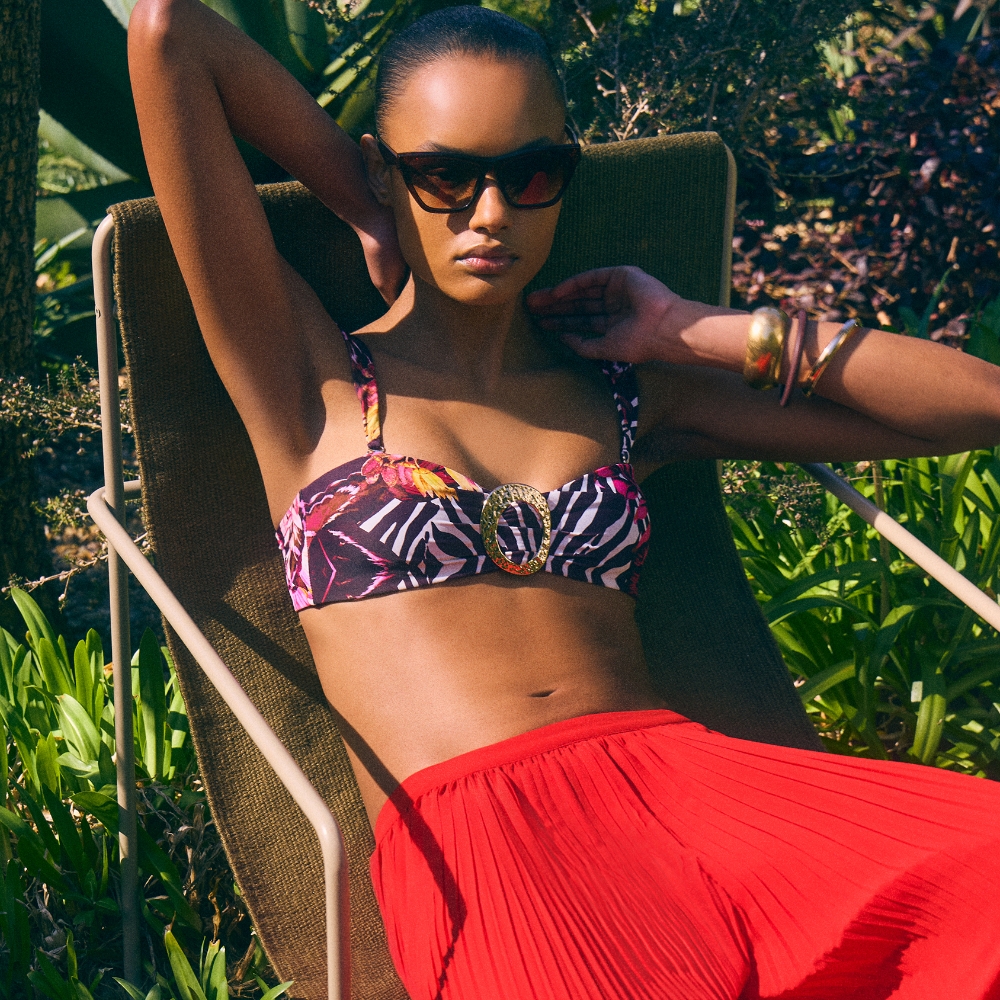 Model wearing a patterned bikini top and red plisse trousers sold at Karen Millen sat on a sun lounger