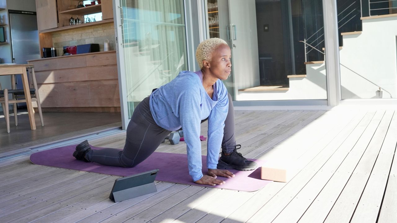 woman on a terrace performing a deep lunge on an exercise mat in front of sunlight to the side of the camera.