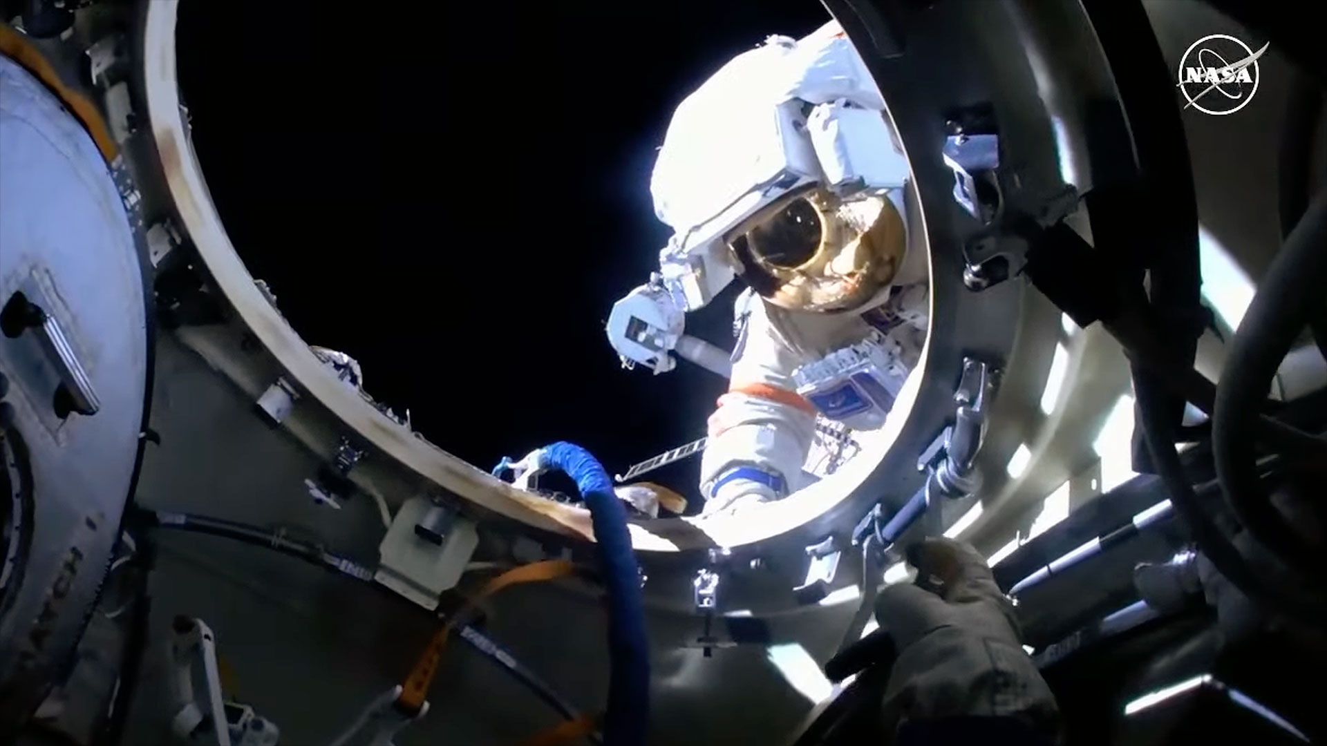 a cosmonaut in a white spacesuit during a spacewalk, as seen through a metallic circular part of the international space station