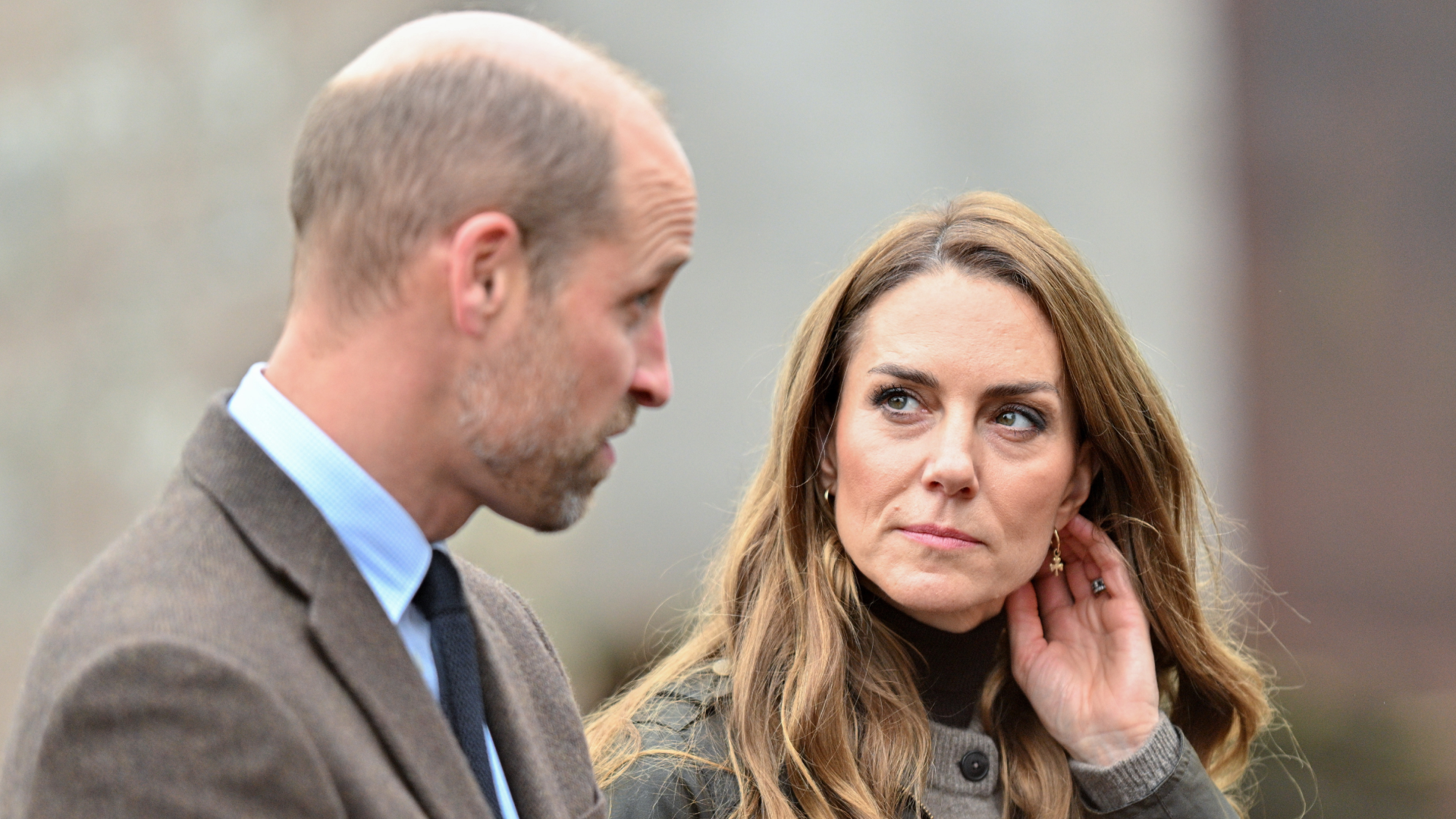 Prince William wearing a brown blazer looking at Princess Kate, putting her hand to her head