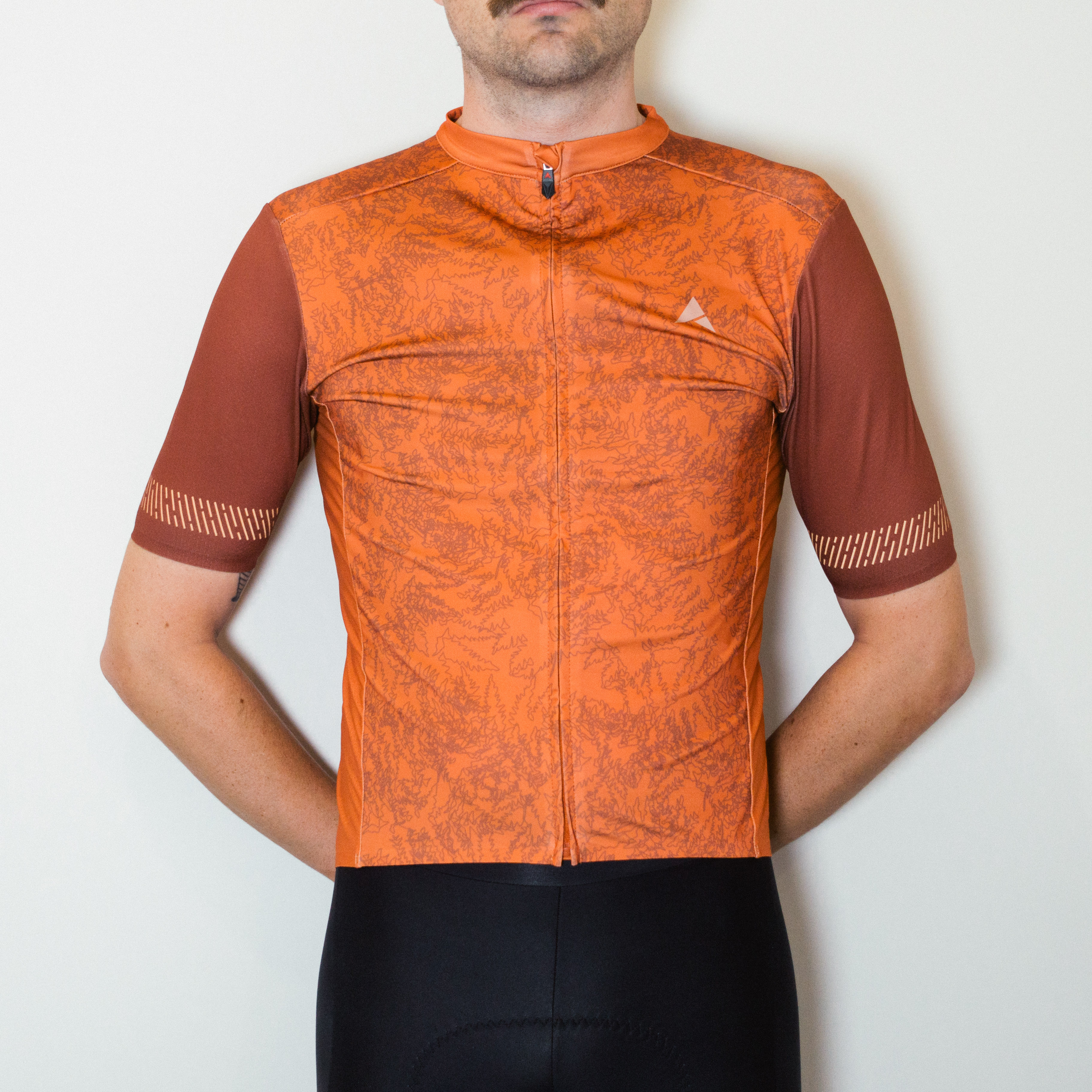 A white man in an orange cycling jersey and black cycling shorts stands against a white wall