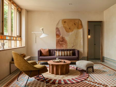 Living space with neutral walls and vibrant, tiled flooring and skirting. An earth toned wall tapestry hangs at the back of the space, with a plum coloured sofa in front. In the foreground sits a round, terracotta coffee table with a mustard armchair and beige, textured footstool surrounding. 