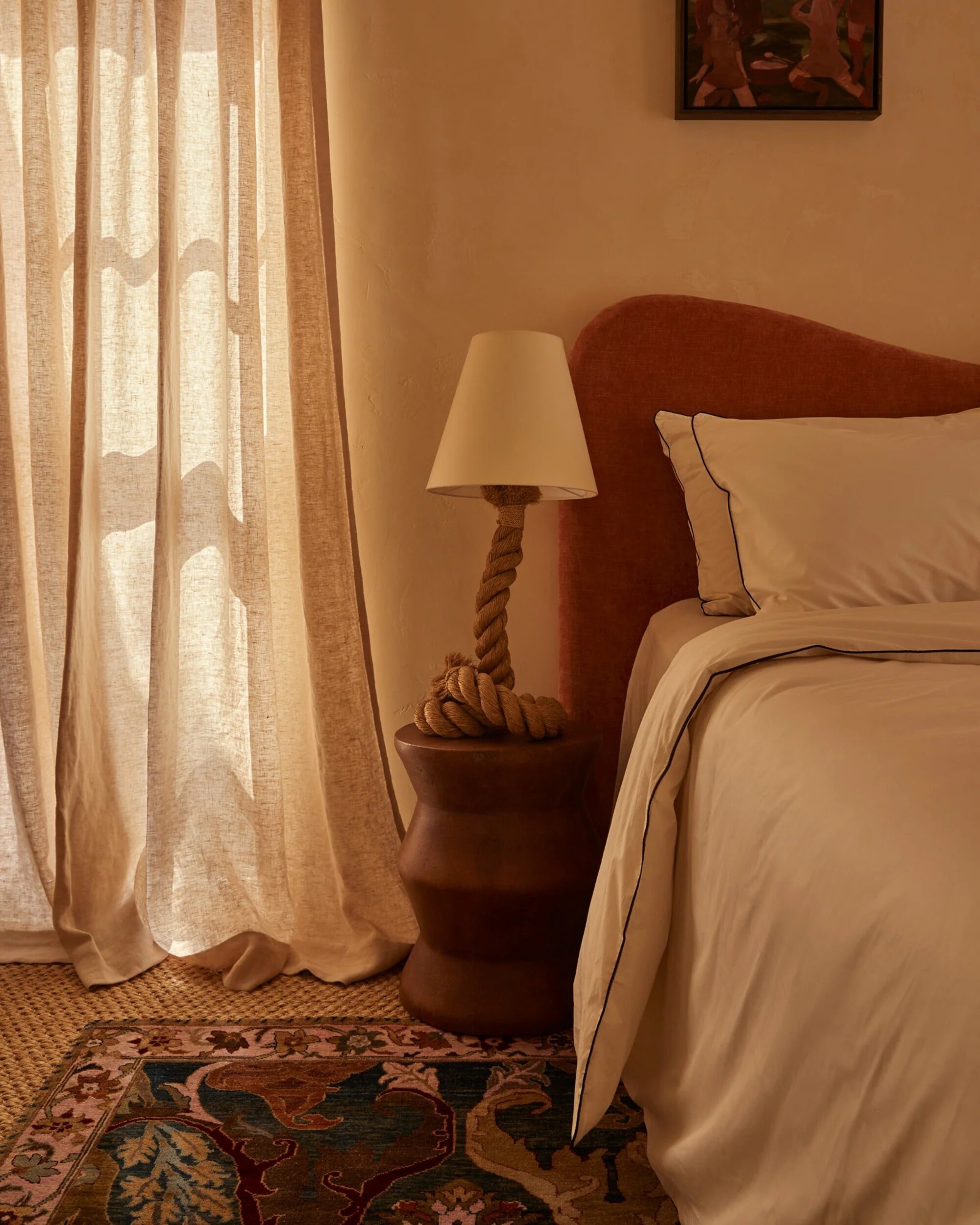 Image of a bedroom with a red bed and light bedding, a wooden side table with a rope table lamp, and a window with relaxed linen curtains.