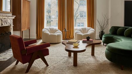 A living room with yellow floor-length curtains against two windows, a green curved sofa, three armchairs, with a sculptural coffee table and freestanding storage unit in a corner