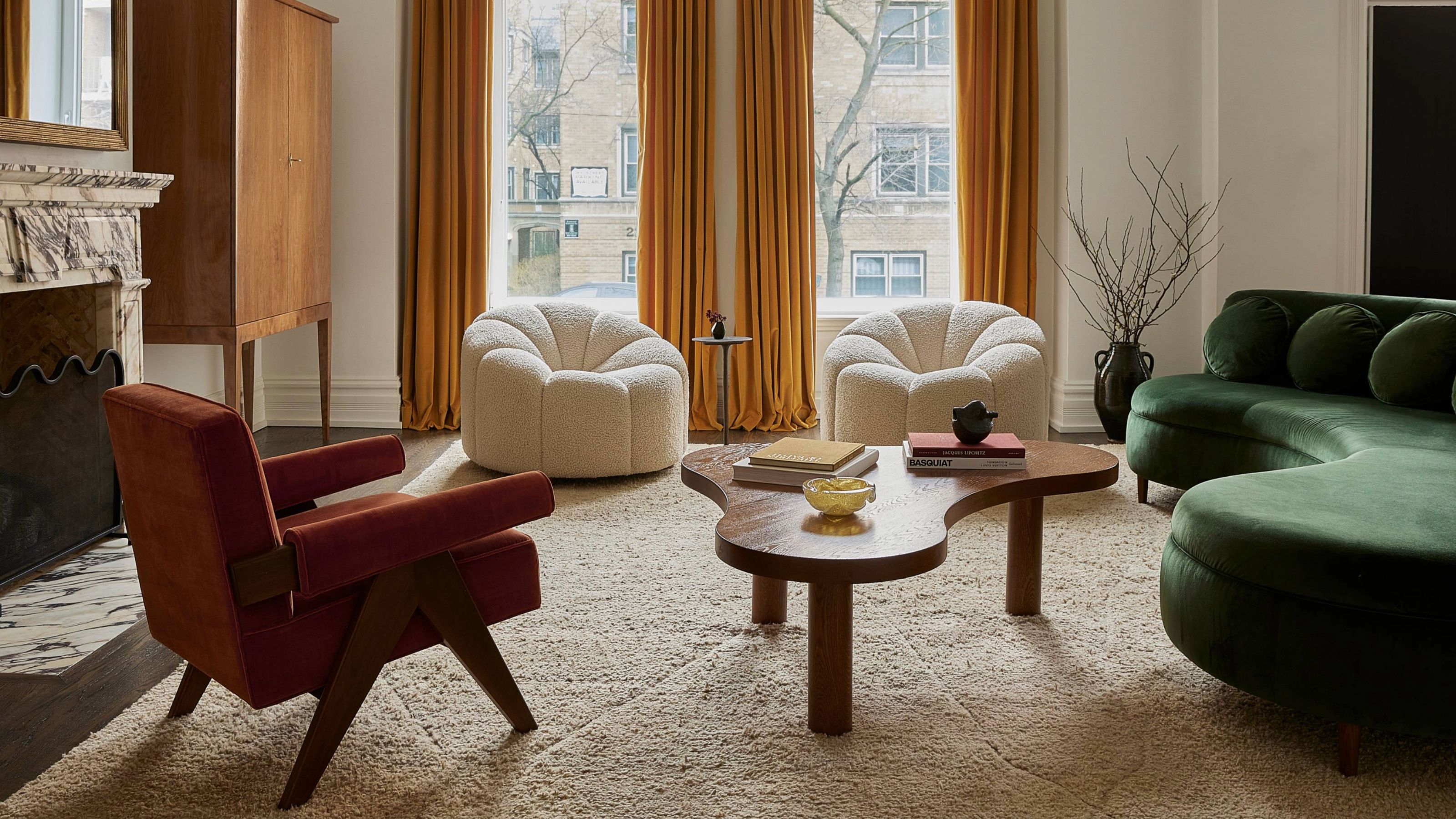A living room with yellow floor-length curtains against two windows, a green curved sofa, three armchairs, with a sculptural coffee table and freestanding storage unit in a corner