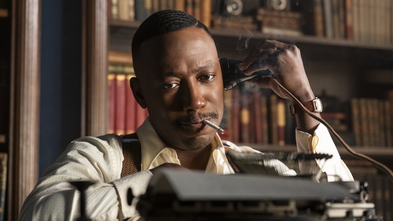 Lamorne Morris as Robbie Robertson seated in front of typewriter and holding old-timey audio speaker to his ear in Spider-Noir