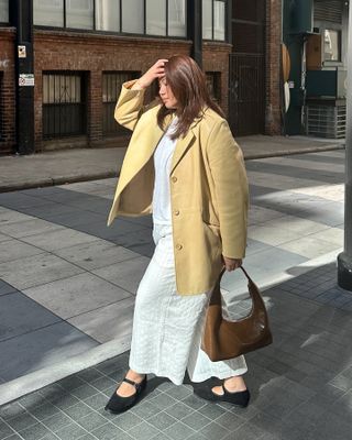 style influencer Marina Torres poses in a butter yellow blazer, a brown patent shoulder bag, white linen pants, and black Mary Jane flats