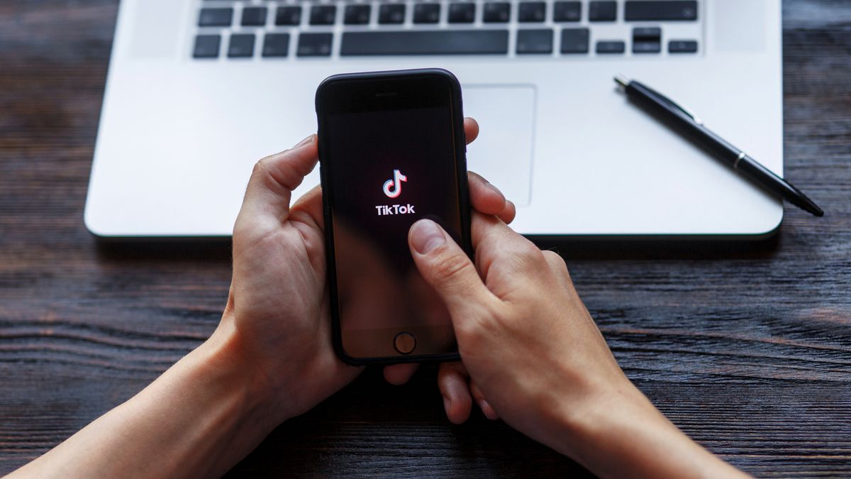 Hands holding a smartphone displaying TikTok logo with laptop in background