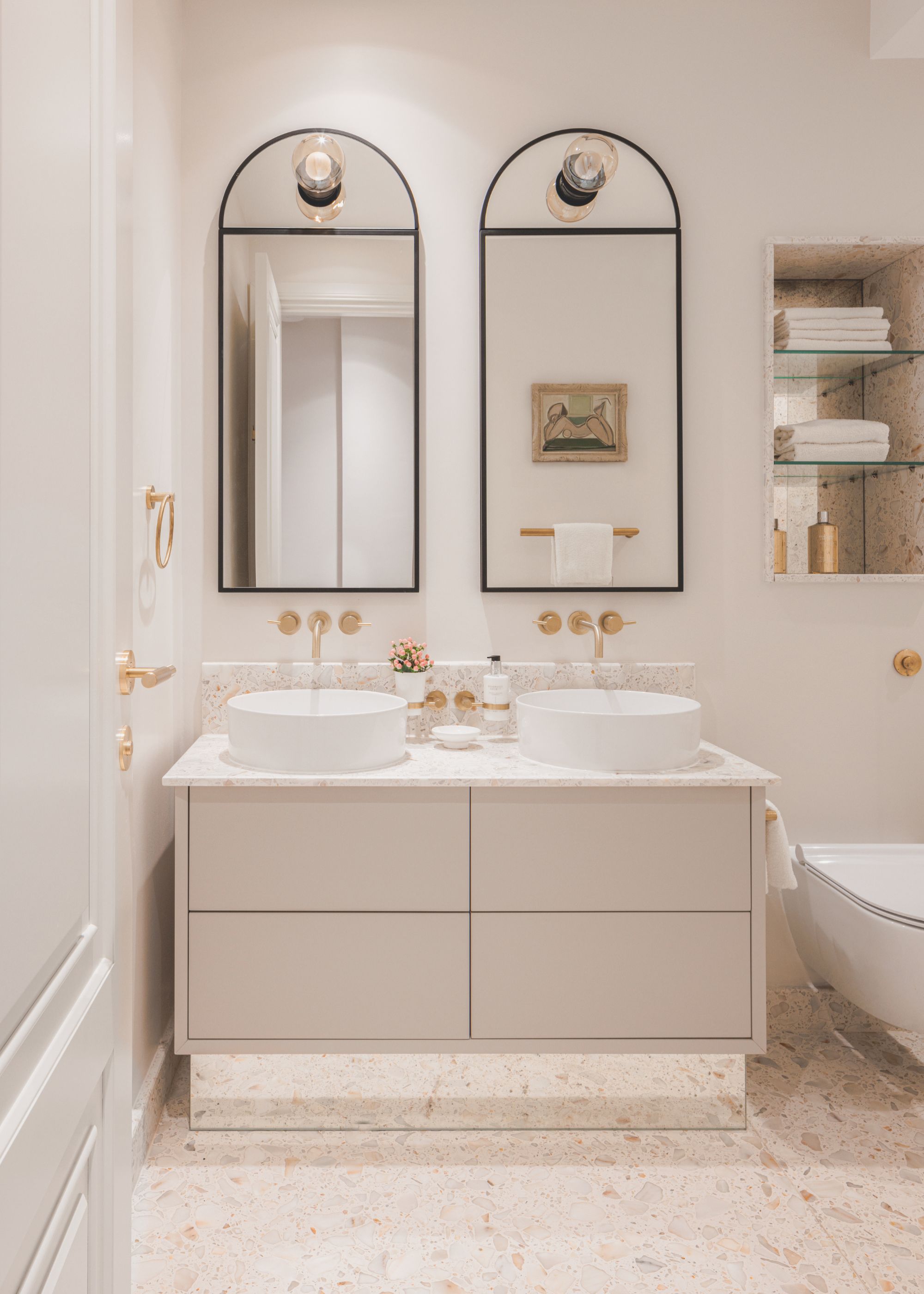 A bathroom with two mirrors and a double basin vanity with gold fixtures above it. On the mirrors are also two large wall lights. There is also a view of open glass shelving above the toiler with towels on top