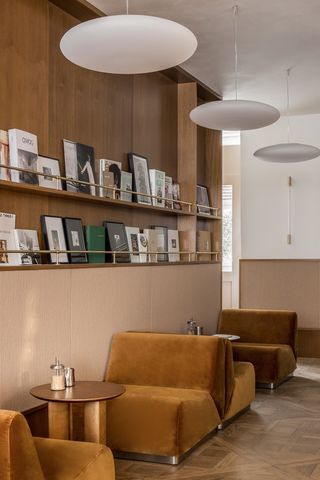 A 1970s-inspired café features velvety love seats in brown, sculptural pendant lighting, and a selection of coffee table books, all stacked in a wall shelf created in a wooden wall.