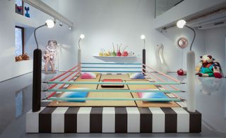 A boxing ring style installation displayed at R & Company’s new New York gallery space. The ring has a zebra style black and white base with pride colour barriers and cushions inside the ring. Lamps on each of the four posts illuminate the display..
