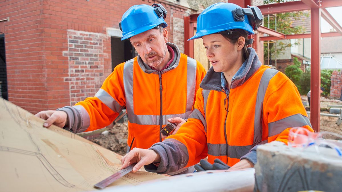 male and female wearing orange hi vis jackets and blue hard hats stood on building site looking at plans