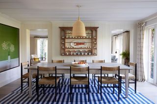 A dining room with wood panelled walls painted in a warm neutral, a blue and white rug and a farmhouse-style dining table