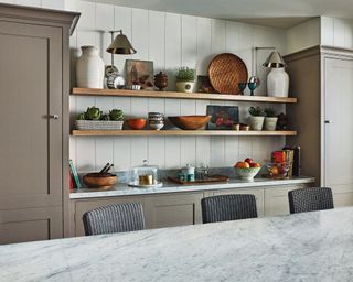 View across a marble topped kitchen island with shelves in the background with dishes and baskets, taupe painted kitchen cupboards