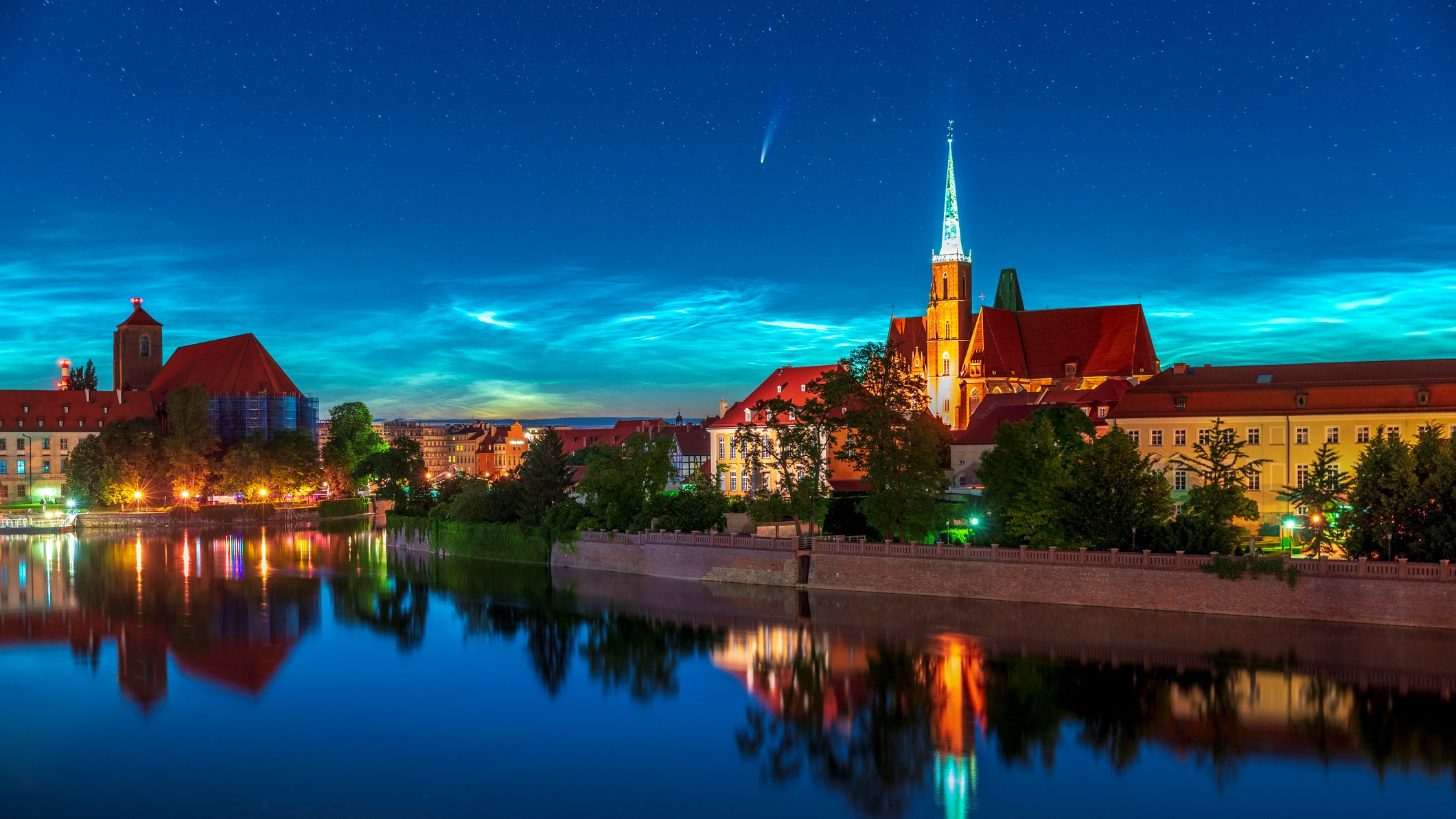 Wispy white clouds shine at twilight, comet NEOWISE streaks across the sky.  A church and trees are reflected in the water below.