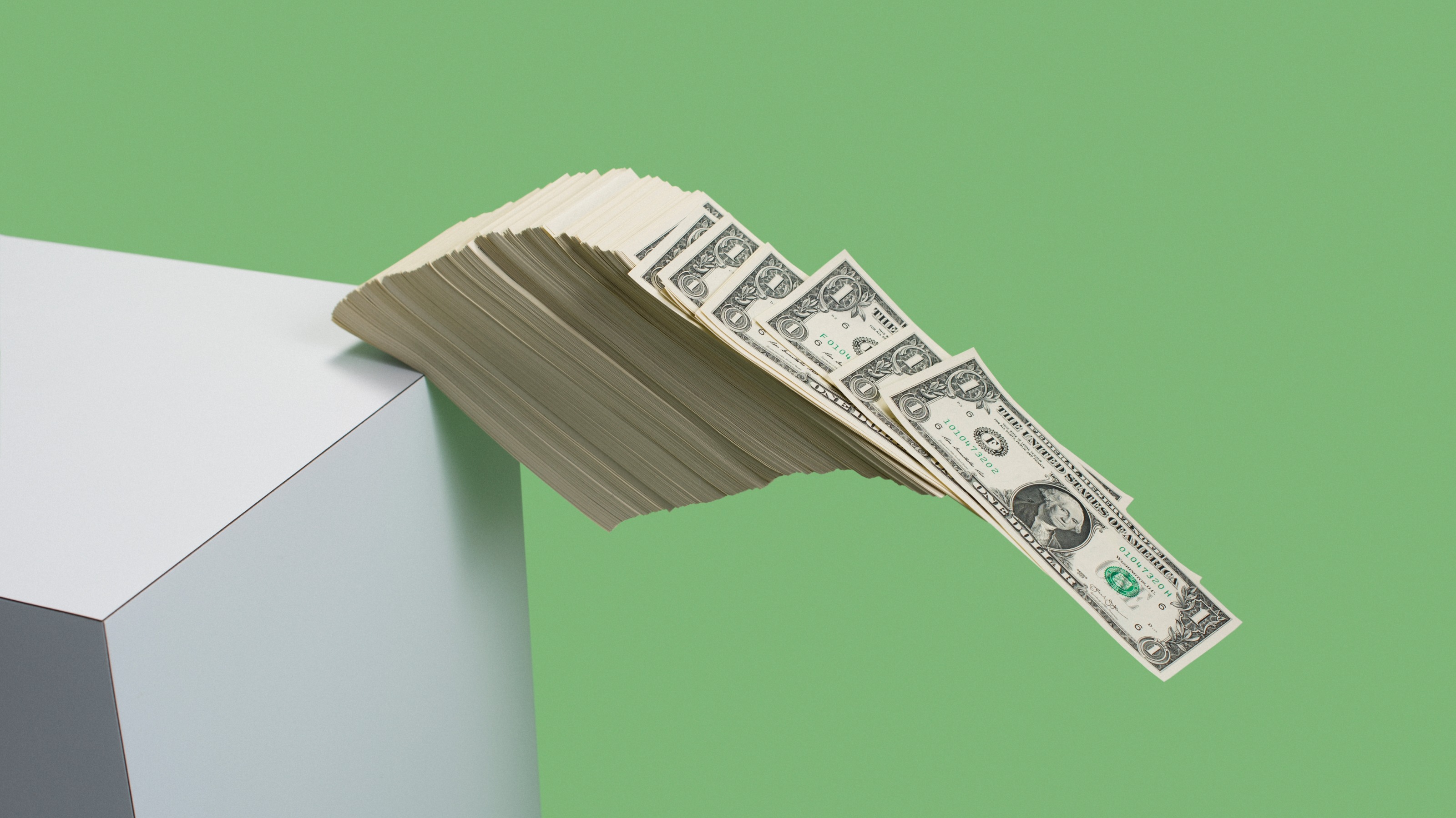 Stack of US $1 bills fall over edge of white shelf on green background